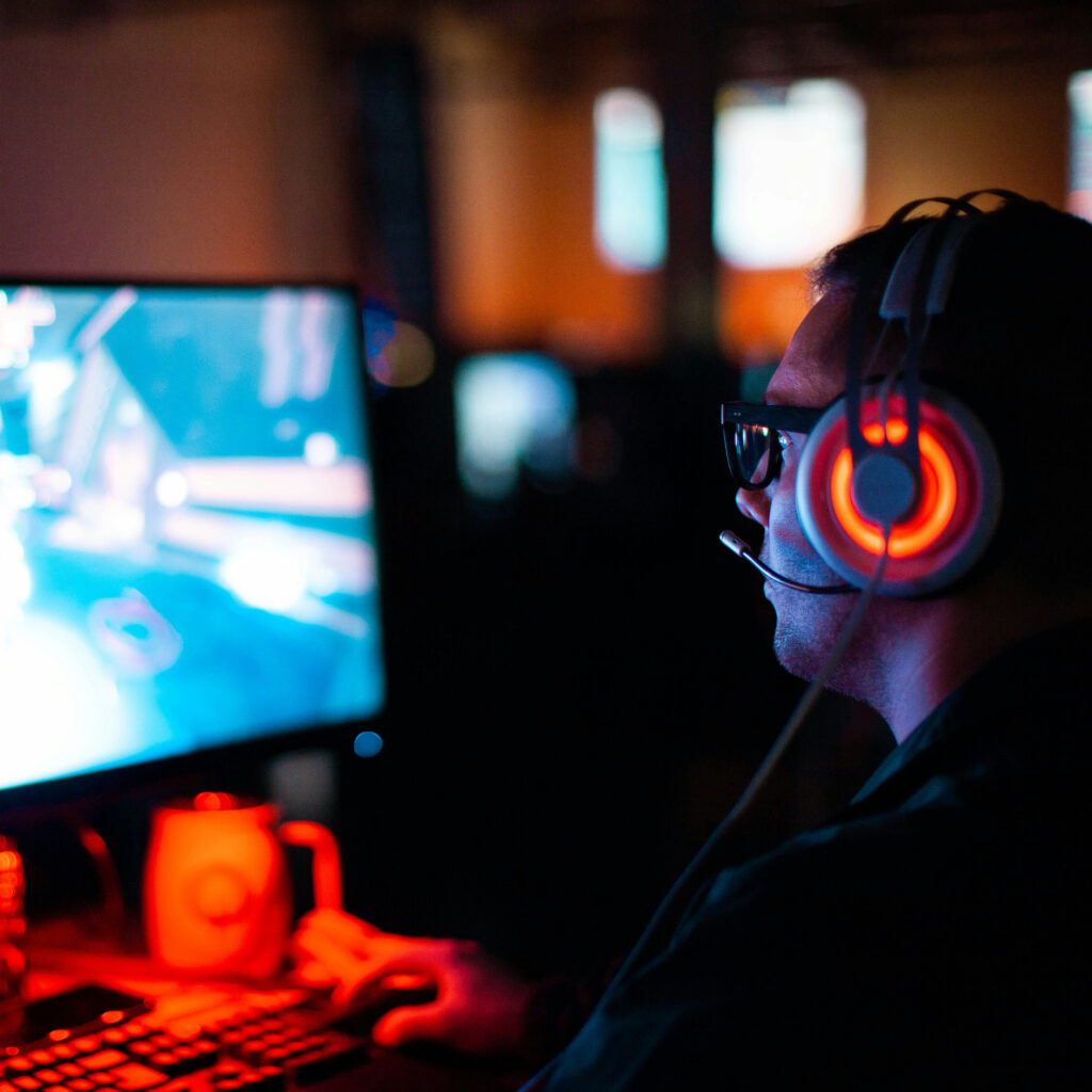 ung man med hörlurar i ett mörkt rum, sitter framför datorskärm och spelar datorspel.